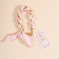 The Darling Effect Sugar Dot Call Me Later Phone Scarf: a chic pink and white polka dot braided wrist strap with gold-tone chain, attached to a pink-edged tag, displayed on a beige surface.