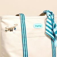 The Darling Effect's Sicily Stripes Teal The Darling Canvas Tote Bag features comfortable teal striped handles, a teal and white scarf on one handle, a gold charm-and-pearl pin on the front pocket, and a roomy interior for essentials.
