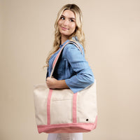 A woman with long blonde hair in a blue denim shirt holds The Darling Effect’s Blush The Signature Canvas Tote Bag, featuring pink straps and durable handles, over her shoulder against a beige background—perfect for everyday use.