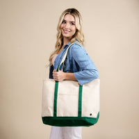 A person with long blonde hair, wearing a light blue denim shirt and white pants, smiles while holding The Darling Effect's Dark Green The Signature Canvas Tote Bag in front of a beige background.