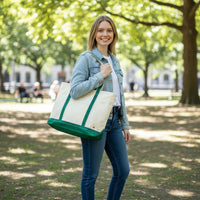 A woman stands outdoors in a sunny park, smiling and holding The Darling Effect's Dark Green The Signature Canvas Tote Bag—featuring green accents and durable handles. She wears a denim jacket, white shirt, and jeans. Trees and people on benches are in the background.