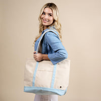 A smiling woman with long blonde hair, in a light blue shirt, carries The Darling Effect's Light Blue The Signature Canvas Tote Bag—featuring durable handles—over her shoulder against a beige background. Perfect for everyday use.