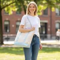 A blonde woman in a white t-shirt and blue jeans walks across a lawn, carrying The Darling Effect’s Light Blue The Signature Canvas Tote Bag with durable handles, perfect for everyday use.