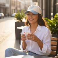 A woman in a white shirt and The Darling Effect's In My Bride Era Classic Canvas Hat sits at an outdoor café table, smiling with coffee in hand and city views behind her, showcasing a relaxed, everyday look.