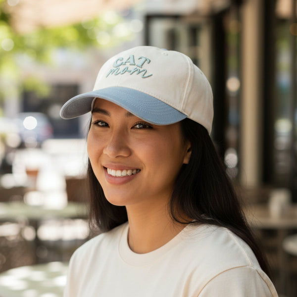 A person beams outdoors, sporting The Darling Effect’s Cat Mom Classic Canvas Hat—crafted from light-colored canvas with “CAT MOM” embroidery—and a cream, relaxed-fit shirt, effortlessly highlighting a laid-back, casual style against a softly blurred backdrop of tables and chairs.