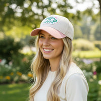 A smiling woman with long blonde hair wears The Darling Effect’s Queen of the Green Classic Canvas Hat in light pink and white, standing outdoors in a sunlit garden with green trees and colorful flowers.