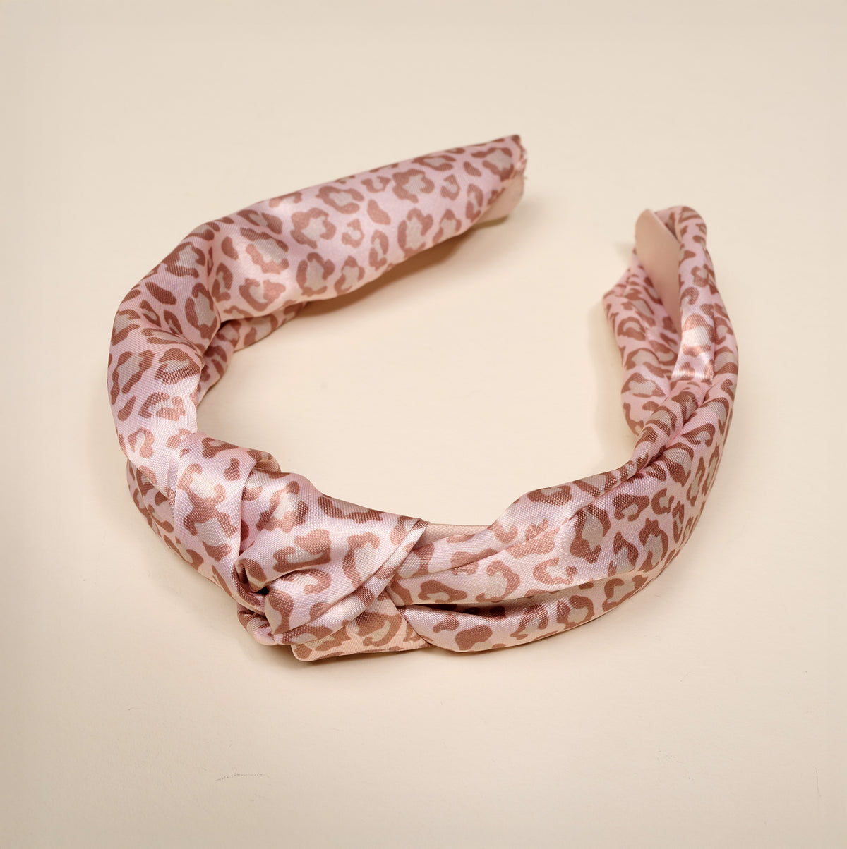 The Born Wild Classic Twist Headband by The Darling Effect features an elegant pink satin design with a subtle leopard print pattern, beautifully displayed on a plain cream-colored background. This versatile hair accessory effortlessly complements any look.