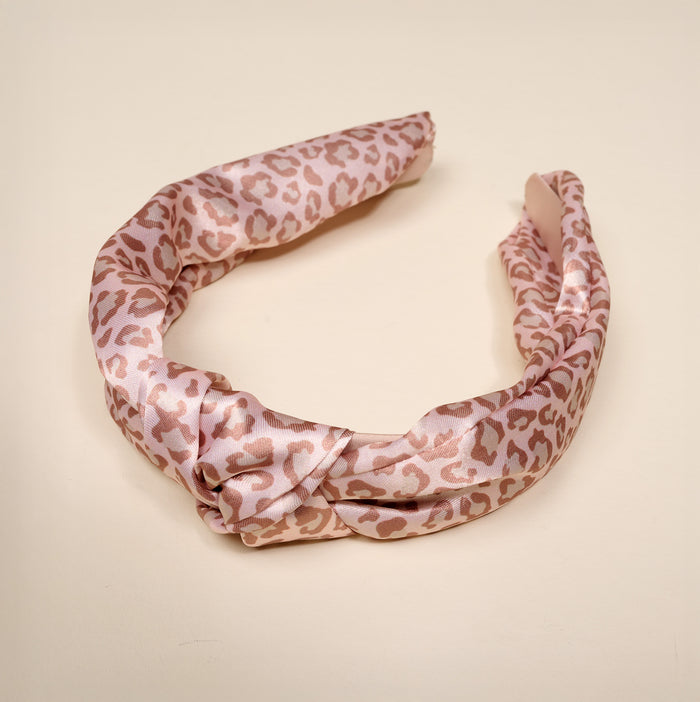 The Born Wild Classic Twist Headband by The Darling Effect features an elegant pink satin design with a subtle leopard print pattern, beautifully displayed on a plain cream-colored background. This versatile hair accessory effortlessly complements any look.