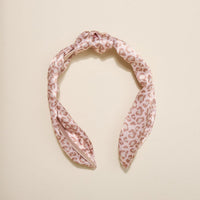 The Darling Effect's Born Wild Classic Twist Headband features a pink satin twist with knot detail and light brown leopard print, shown on a beige background—a trendy accessory for any style.