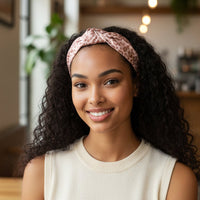 A young woman with long curly hair wears The Darling Effect’s Born Wild Classic Twist Headband, featuring pink leopard print and a knot detail, as she smiles indoors in soft light against a blurred background.