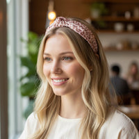 A young woman with long blonde hair smiles in a cozy café, wearing The Darling Effect's Born Wild Classic Twist Headband—a pink leopard-print accessory with a chic knot detail. Blurred plants and shelves fill the softly lit background.