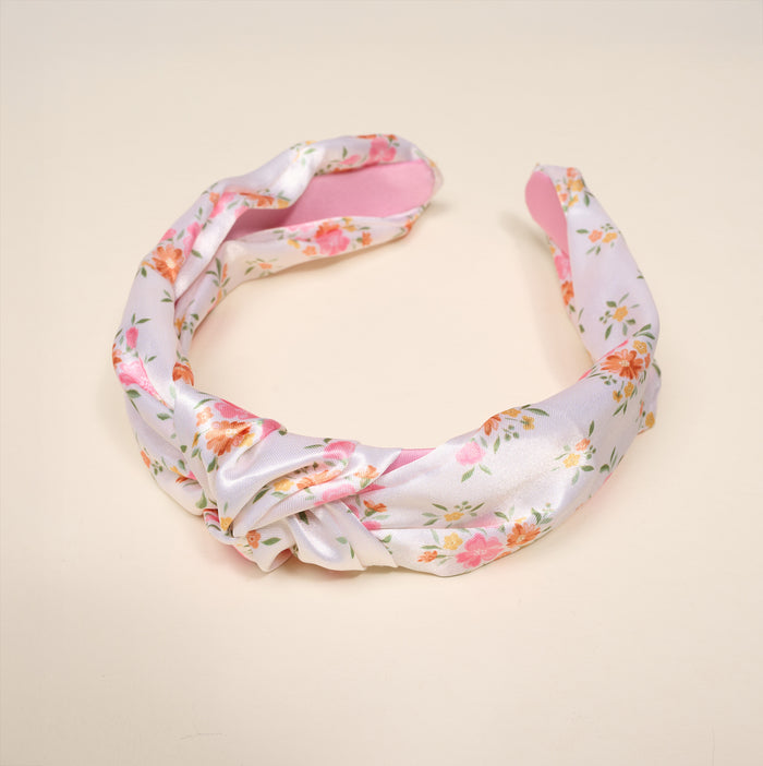 The Darling Effect’s Endless Daydream Cream Classic Twist Headband, featuring a pink and white floral satin design with an elegant front knot, is showcased against a light beige background.