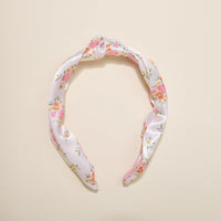 The Darling Effect’s Endless Daydream Cream Classic Twist Headband features a white satin finish with pink, orange, and green floral patterns—an elegant, versatile accessory for any occasion, shown on a beige background.