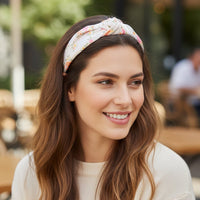 A woman with long brown hair smiles while sitting outdoors at a café, wearing The Darling Effect's Endless Daydream Cream Classic Twist Headband and a cream-colored sweater.