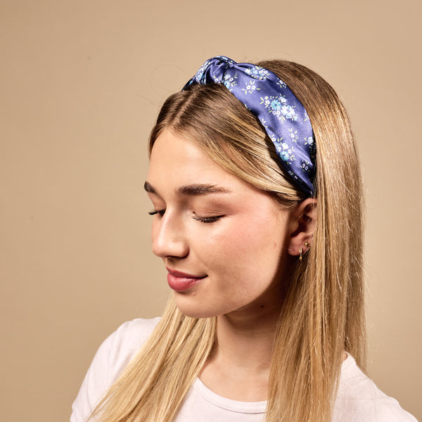 A young woman with long blonde hair wears The Darling Effect's Endless Daydream Navy Classic Twist Headband and a white shirt, smiling softly as she looks down against a plain beige background.