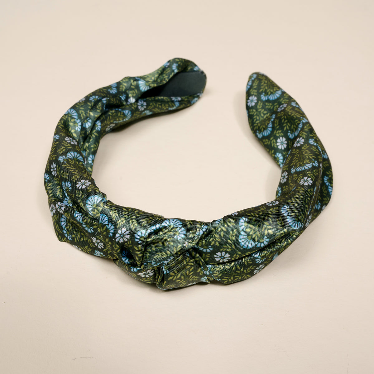 The Everbloom Forest Green Classic Twist Headband by The Darling Effect is a dark green accessory adorned with a blue and white floral pattern. Twisted into a circular shape, it is displayed on a plain light beige background. This versatile headband adds both style and comfort to any look.