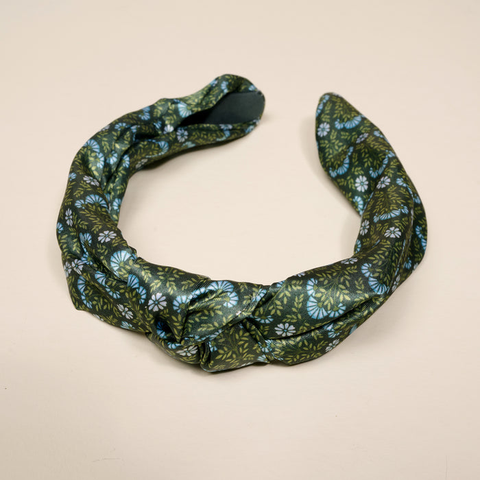 The Everbloom Forest Green Classic Twist Headband by The Darling Effect is a dark green accessory adorned with a blue and white floral pattern. Twisted into a circular shape, it is displayed on a plain light beige background. This versatile headband adds both style and comfort to any look.