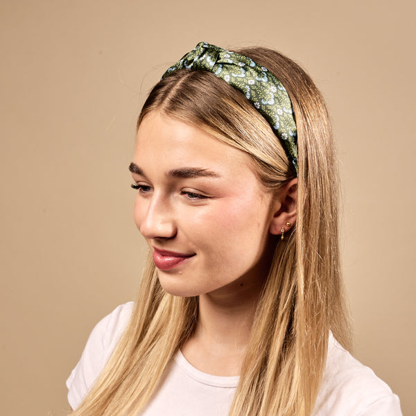 A young woman with straight blonde hair wears The Darling Effect's Everbloom Forest Green Classic Twist Headband and a white t-shirt, smiling softly while looking slightly to the side against a beige background.
