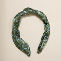The Darling Effect Everbloom Forest Green Classic Twist Headband, featuring a white and blue floral pattern and a stylish knot detail, is displayed against a plain beige background.