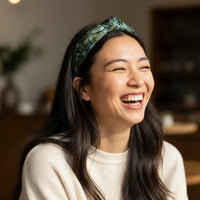 A young woman with long dark hair wears The Darling Effect's Everbloom Forest Green Classic Twist Headband and a cream sweater, smiling and laughing indoors against a blurred background.