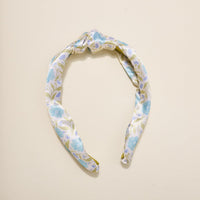 The Darling Effect’s Poppin' Petals Aqua Classic Twist Headband, featuring a blue floral pattern and elegant top knot, rests on a cream background—making it a charming hair accessory.