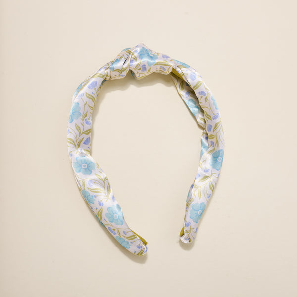 The Darling Effect’s Poppin' Petals Aqua Classic Twist Headband, featuring a blue floral pattern and elegant top knot, rests on a cream background—making it a charming hair accessory.