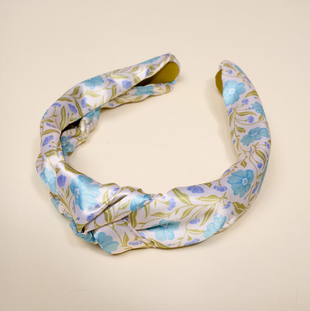 The Darling Effect’s Poppin’ Petals Aqua Classic Twist Headband showcases a satin knot design with a floral pattern in blue and green hues on a light beige background, displayed on a neutral surface. This versatile hair accessory brings an elegant touch to any hairstyle.