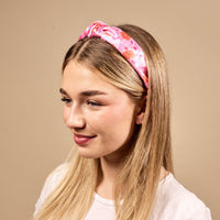 A young woman with long blonde hair smiles slightly and looks to the side, wearing The Darling Effect's Poppin' Petals Hot Pink Classic Twist Headband and a white shirt against a plain beige background.