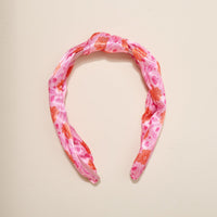 The Darling Effect's Poppin' Petals Hot Pink Classic Twist Headband, featuring bold orange and red floral patterns on pink fabric, is displayed on an off-white background—a stylish accessory for any occasion.