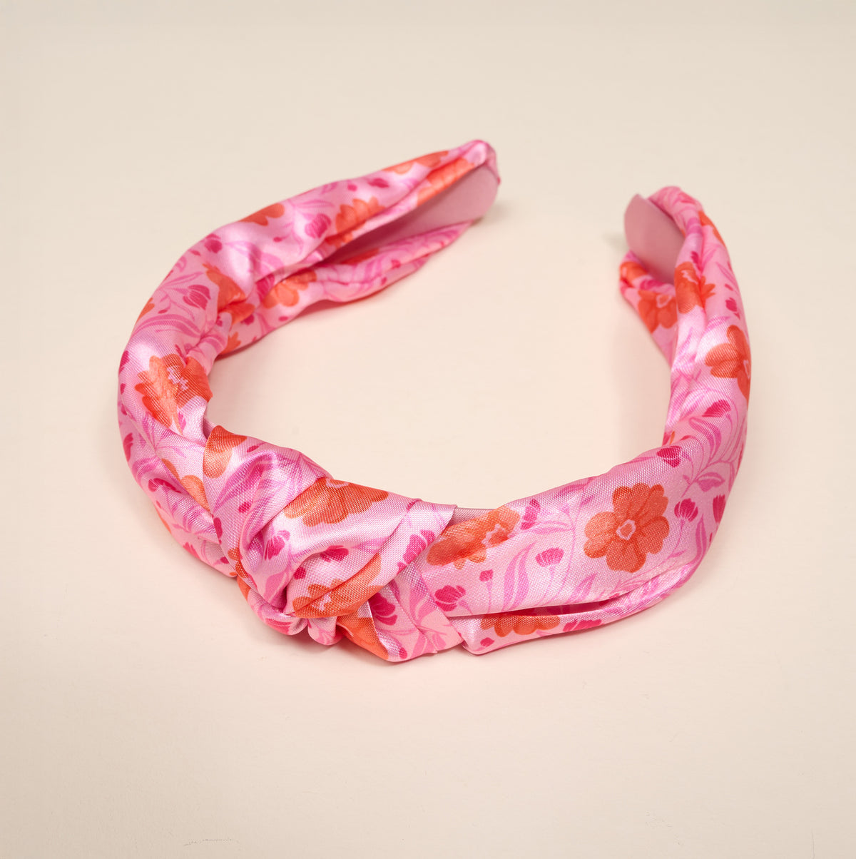 The Darling Effect’s Poppin’ Petals Hot Pink Classic Twist Headband is a satin hair accessory with a knotted center detail, showcasing a vibrant floral pattern in shades of pink and orange. Displayed on a plain light background, this chic headband adds an elegant finishing touch to any outfit.