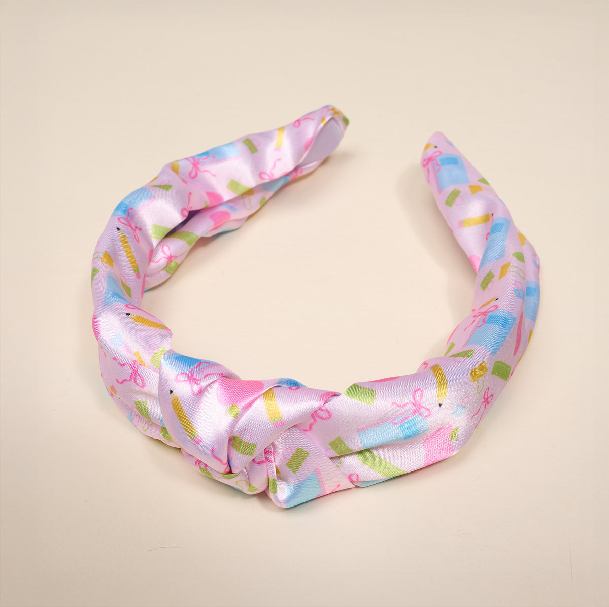 The School’s In Session Classic Twist Headband by The Darling Effect features a pastel pink satin design with a central knot, adorned with colorful book and bow patterns on a plain beige background—making this versatile accessory an effortless way to add charm to any look.