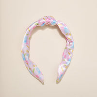 The Darling Effect’s School's In Session Classic Twist Headband features pink satin with a top knot and a vibrant pattern of blue, yellow, and green scissors and rectangles—a fun accessory for any occasion, shown on a plain beige background.