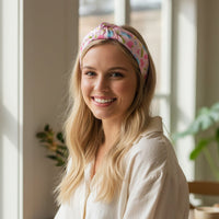 A young woman with long blonde hair smiles while wearing The Darling Effect’s School's In Session Classic Twist Headband and a cream shirt, sitting indoors by a sunny window with green plants in the background.