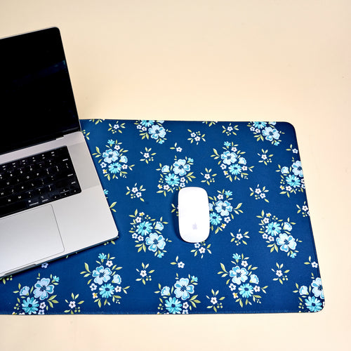 A laptop and a white mouse rest on the-darling-effect’s Endless Daydream Navy Style Station Desk Mat, featuring light blue and white flowers—a stylish workspace accessory atop a beige surface.