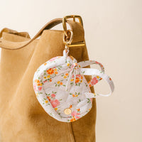 The-darling-effect's Endless Daydream Cream Itty Bitty Duffle Bag Charm is a tan suede accessory with a gold clasp and floral face mask holder, featuring pink and orange flowers on white, attached with a gold keyring.