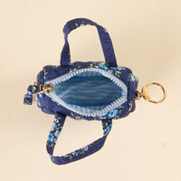 The-darling-effect’s Endless Daydream Navy Itty Bitty Duffle Bag Charm features a small blue floral-quilted pouch with two handles, gold keyring, zipper, and light blue-white striped lining—perfect as a charming bag accessory. Displayed on beige.