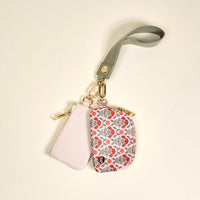 The Darling Effect’s Everbloom Dusty Rose Dynamic Duo Pouch Wristlet features two zippered pouches—one light pink, one white with red and grey florals—hanging from a green wristlet strap and secured with gold clasps for hands-free style.