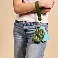 A person in jeans and a white tee holds The Darling Effect's Everbloom Forest Green Dynamic Duo Pouch Wristlet, featuring attached floral green and light blue pouches—perfect for keeping essentials hands-free.