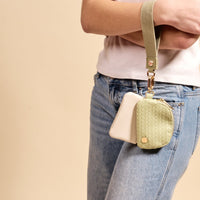 A person carries The Darling Effect’s Sage Dynamic Duo Pouch Luxe Wristlet with pale green and white pouches and gold hardware, while wearing light blue jeans and a white shirt against a beige background.