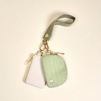 The Darling Effect's Sage Dynamic Duo Pouch Luxe Wristlet features two small zippered pouches—white and sage, one with a woven texture—attached to a light green strap with gold hardware, shown on a cream background.