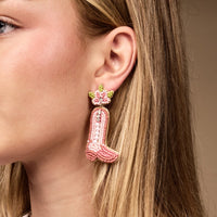 A woman with straight blonde hair wears The Darling Effect’s Cowgirl Boot Beaded Earrings—vibrant pink, hand-beaded with a floral detail. Her face is partially visible against a neutral background.