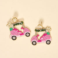 The Darling Effect's Golf Cart Beaded Earrings, handmade in playful pink with white, green, black, and gold accents, are showcased on a light beige background—perfect for fans of colorful accessories.