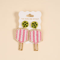 Pickleball Paddle Beaded Earrings by The Darling Effect feature green circular tops, pink-and-white striped paddle-shaped dangles, and gold and white bead accents. Hand-beaded and displayed on a branded card.