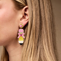 A close-up of a woman wearing The Darling Effect's Pencil Me In Beaded Earrings—colorful pencil-shaped earrings crafted from pink, yellow, white, and black beads—partially hidden by her straight blonde hair.