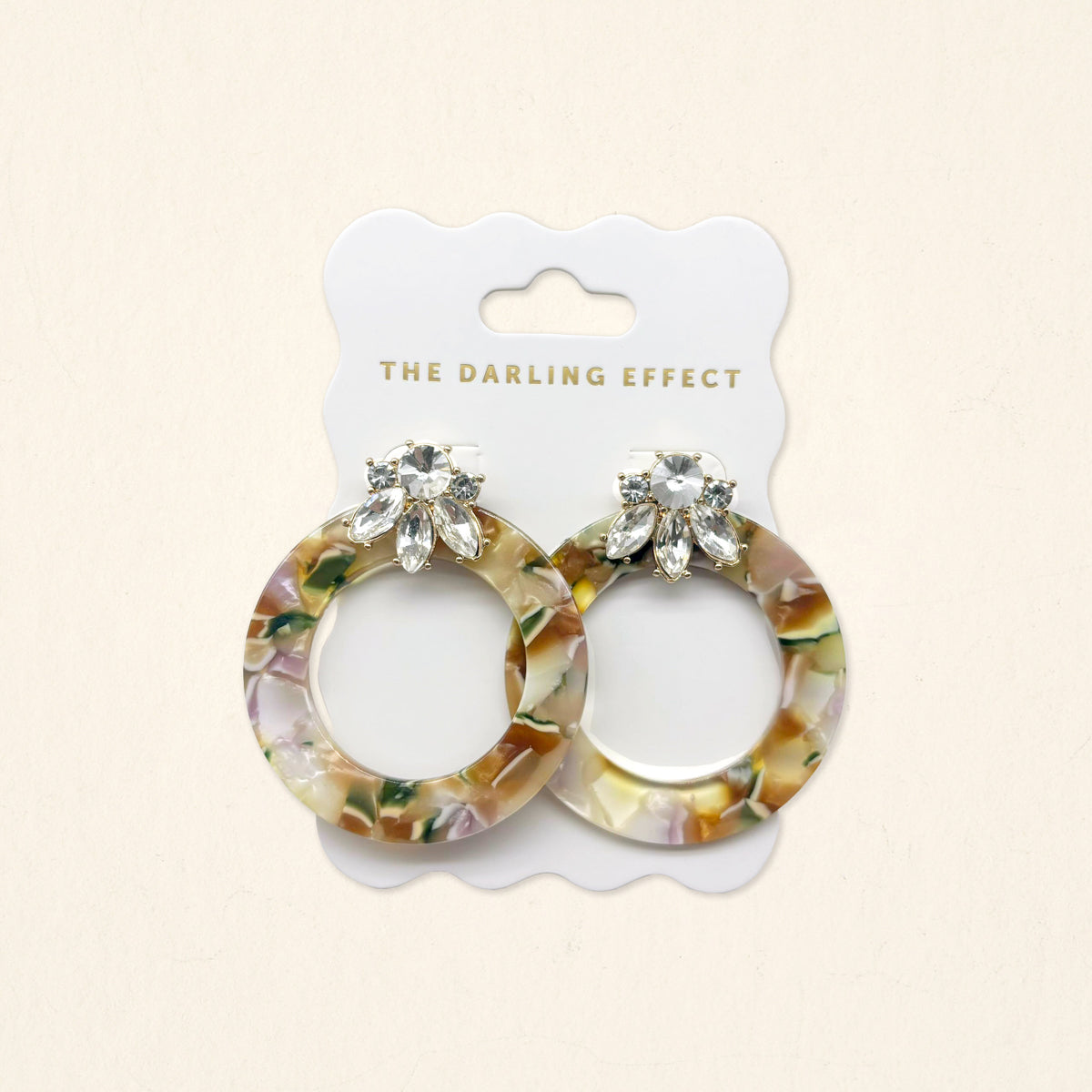 The Amber Drift Glow Getter Earrings by The Darling Effect feature glossy marbled resin, clear crystals, and gold hoops on a white scalloped card with The Darling Effect logo.