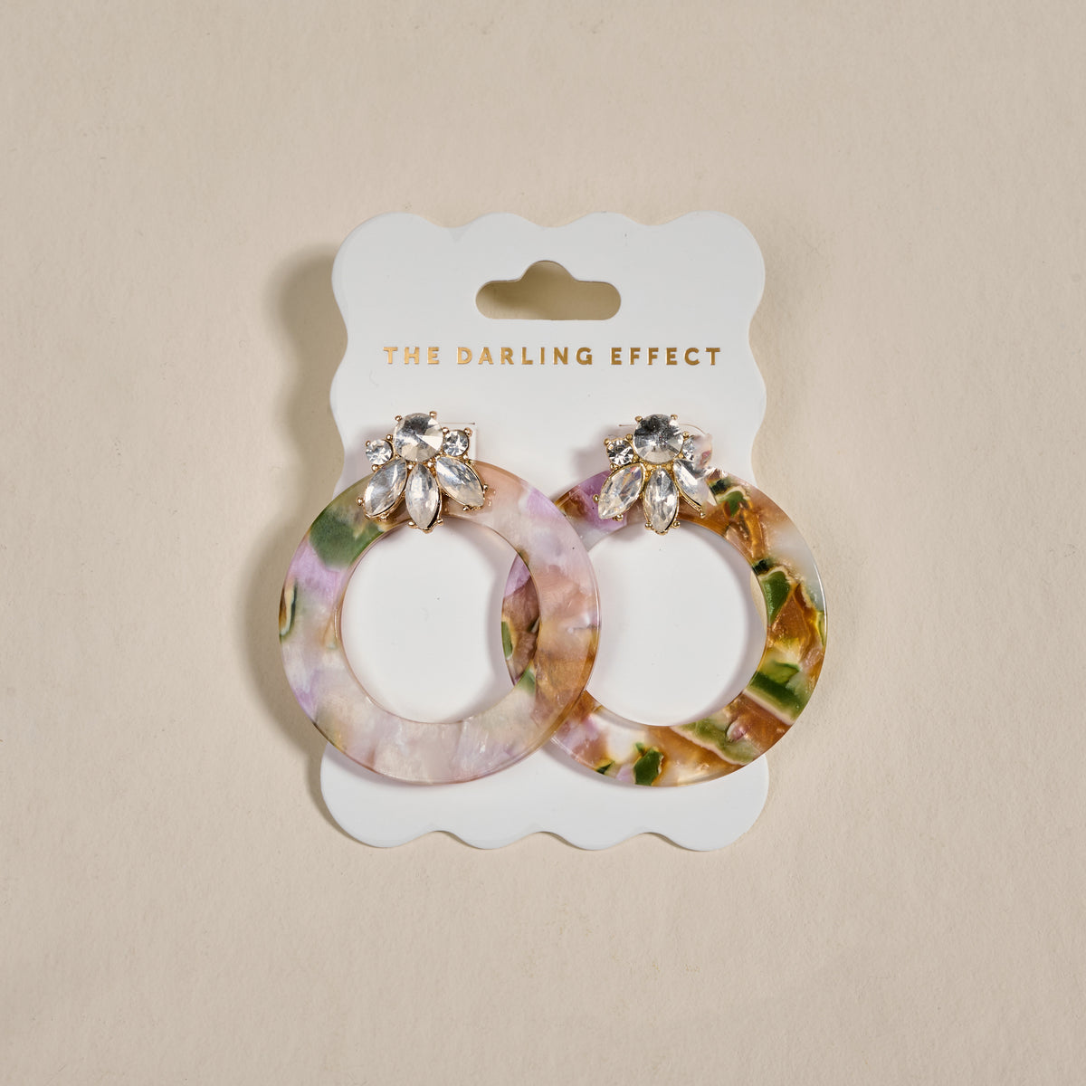 Amber Drift Glow Getter Earrings by The Darling Effect: round, multicolored marbled earrings with rhinestone flower-shaped studs, shown on a white THE DARLING EFFECT card against a beige background.