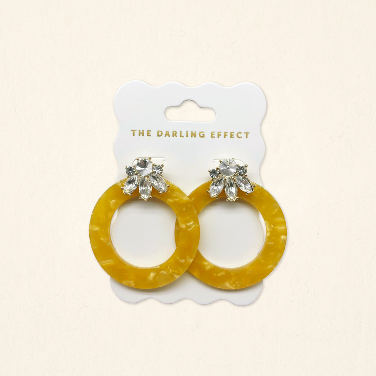 The Darling Effect Honey Gold Glow Getter Earrings feature mustard yellow glossy acetate hoops and crystal rhinestone flower studs on a white scalloped card. Lightweight statement earrings to add a bold, stylish touch to any outfit.