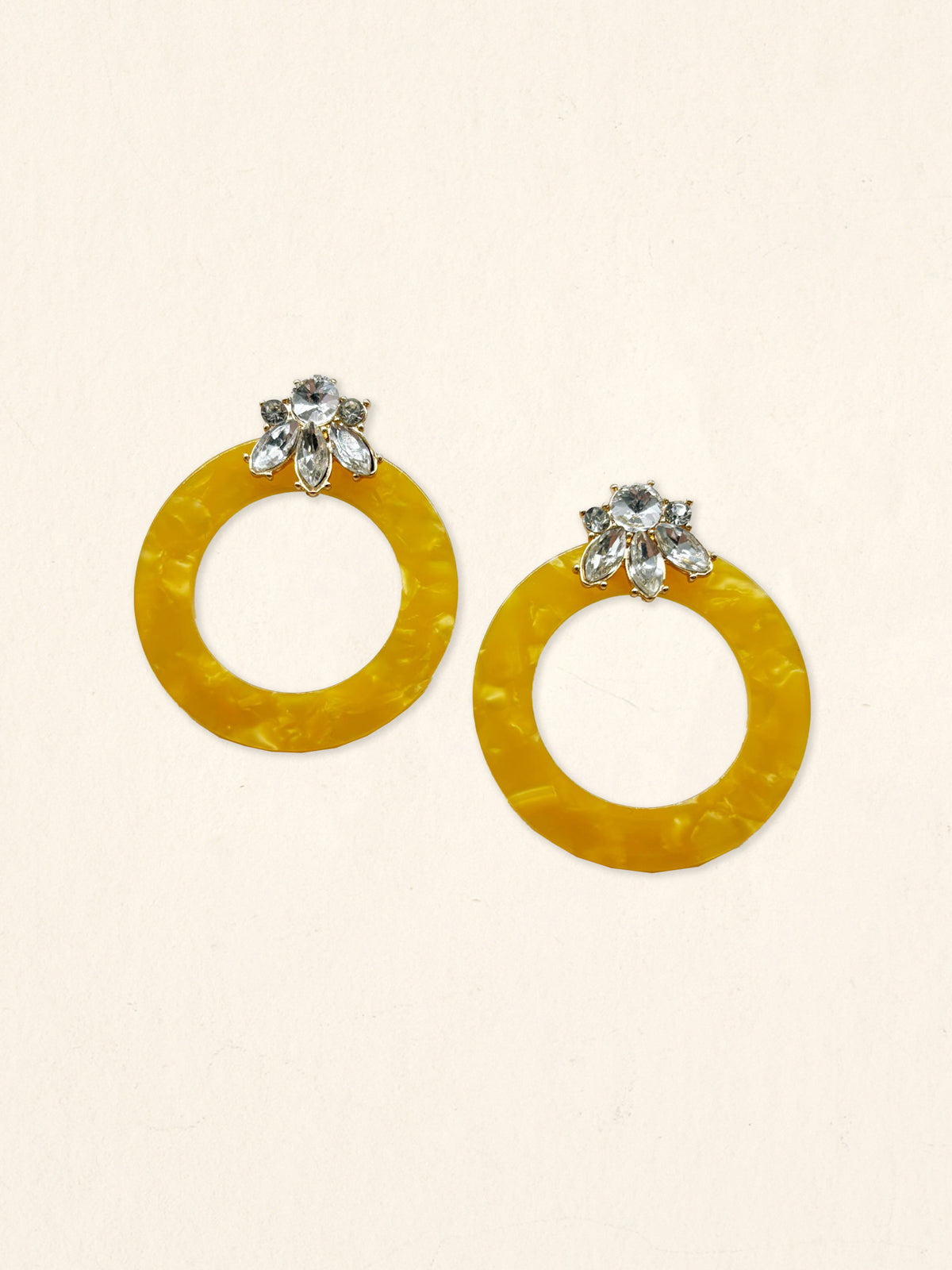 The Darling Effect's Honey Gold Glow Getter Earrings feature large, round yellow hoops and silver floral studs with clear gemstones, shown against a plain beige background.