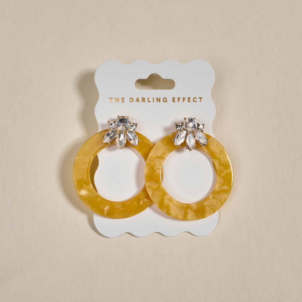 The Darling Effect’s Honey Gold Glow Getter Earrings feature large yellow textured hoops with silver, jewel-encrusted tops, displayed on a white card labeled THE DARLING EFFECT against a neutral background.