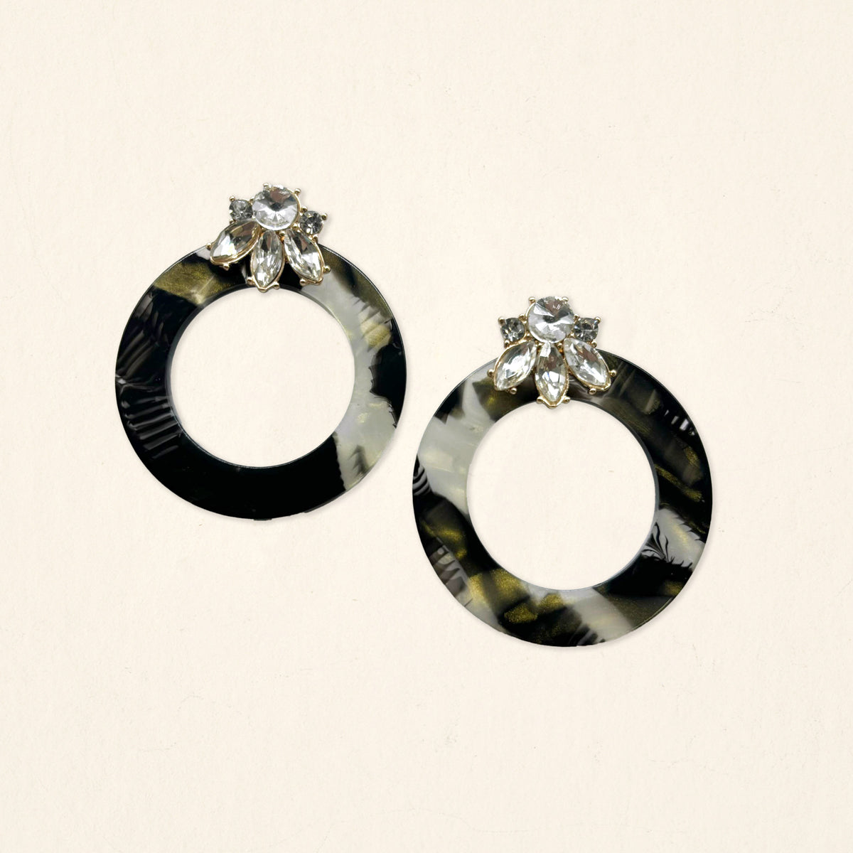 The Darling Effect's Midnight Opal Glow Getter Earrings—round, black and gray marbled with clear flower-shaped gems—are displayed on a light beige background.